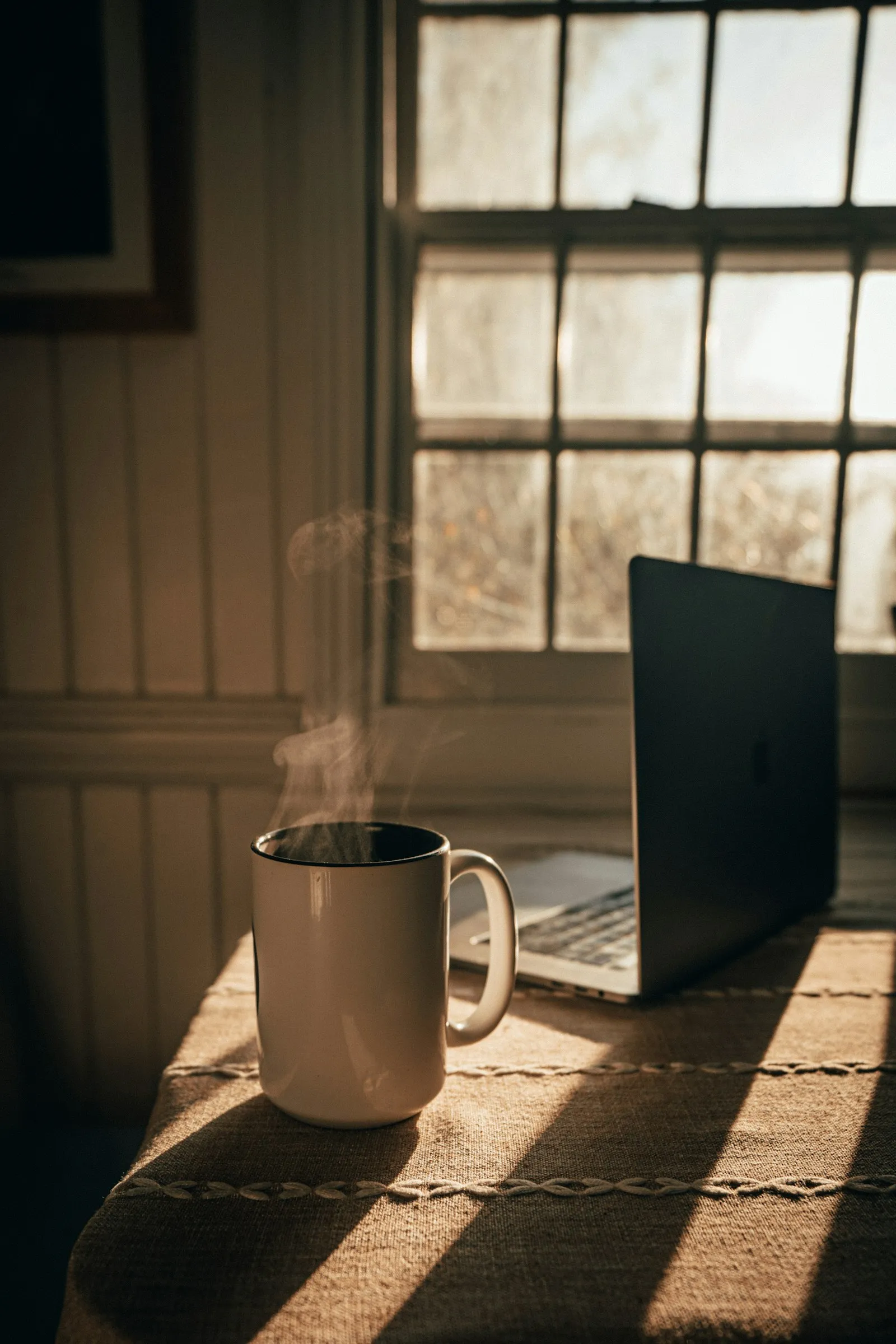 A steaming mug beside an open laptop at a sunny window
